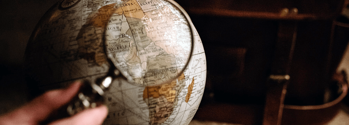 A hand holding a magnifying glass in front of an earth globe with an old leather breafcase in the background.