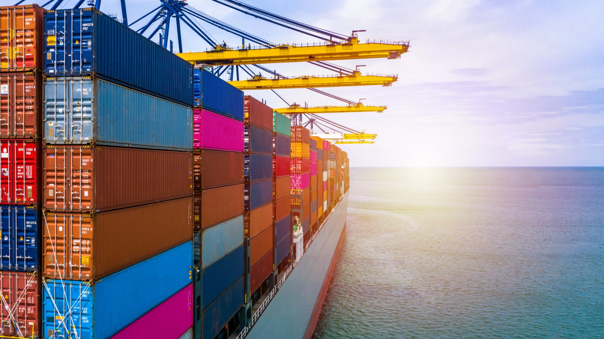 A cargo ship filled with containers in the foreground is positioned under quay cranes, with the ocean stretching out to the horizon and the sun rising and beckoning the container ship to towards its next foray across the open seas