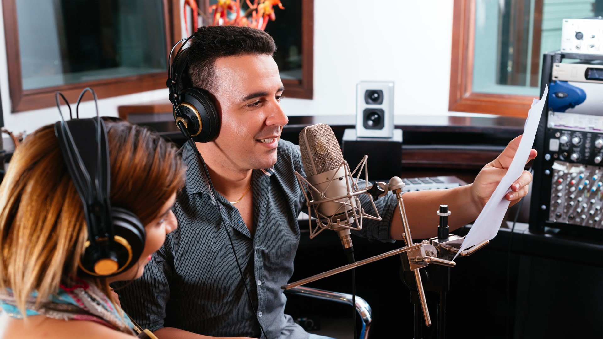 Man and woman are in a recording booth, and the man is reading or singing into a microphone from a paper in front of him. The woman is joining along, or possibly just wondering what the man is doing.