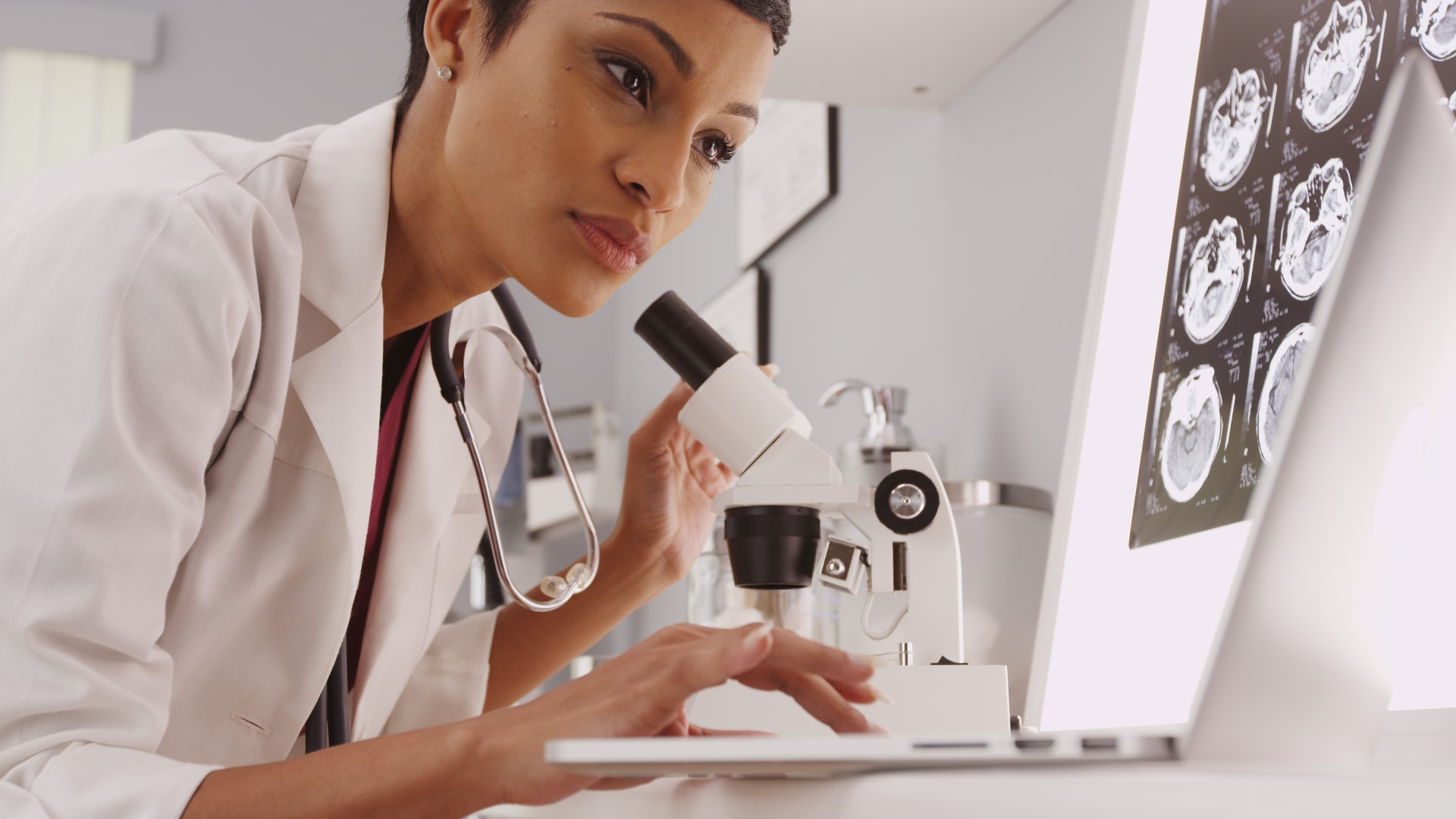 Female doctor with a stethoscope around her neck checks something on her computer after looking through a microscope, with brain scans illuminated on the board in front of her