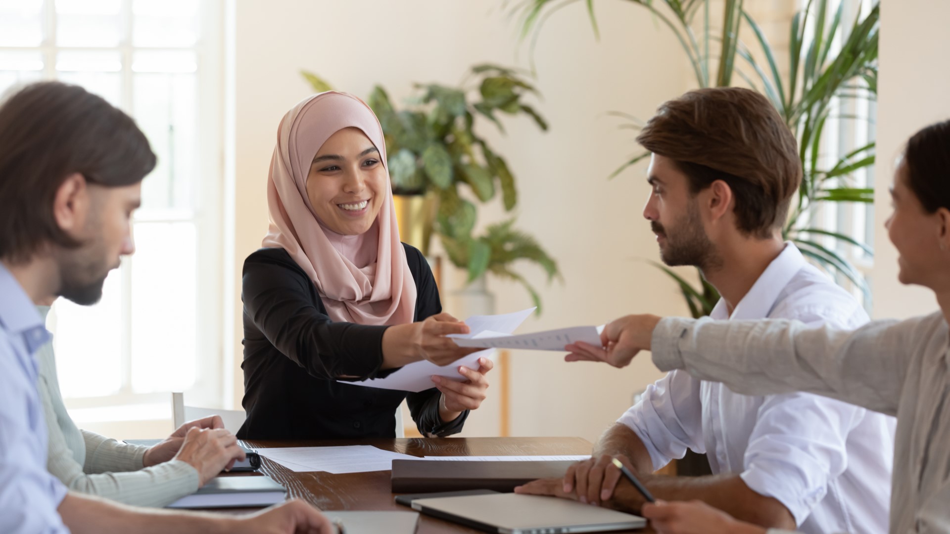 Five employees are sitting at a table with some office plants in the background, and one of the employees hands the woman leading the meeting a document