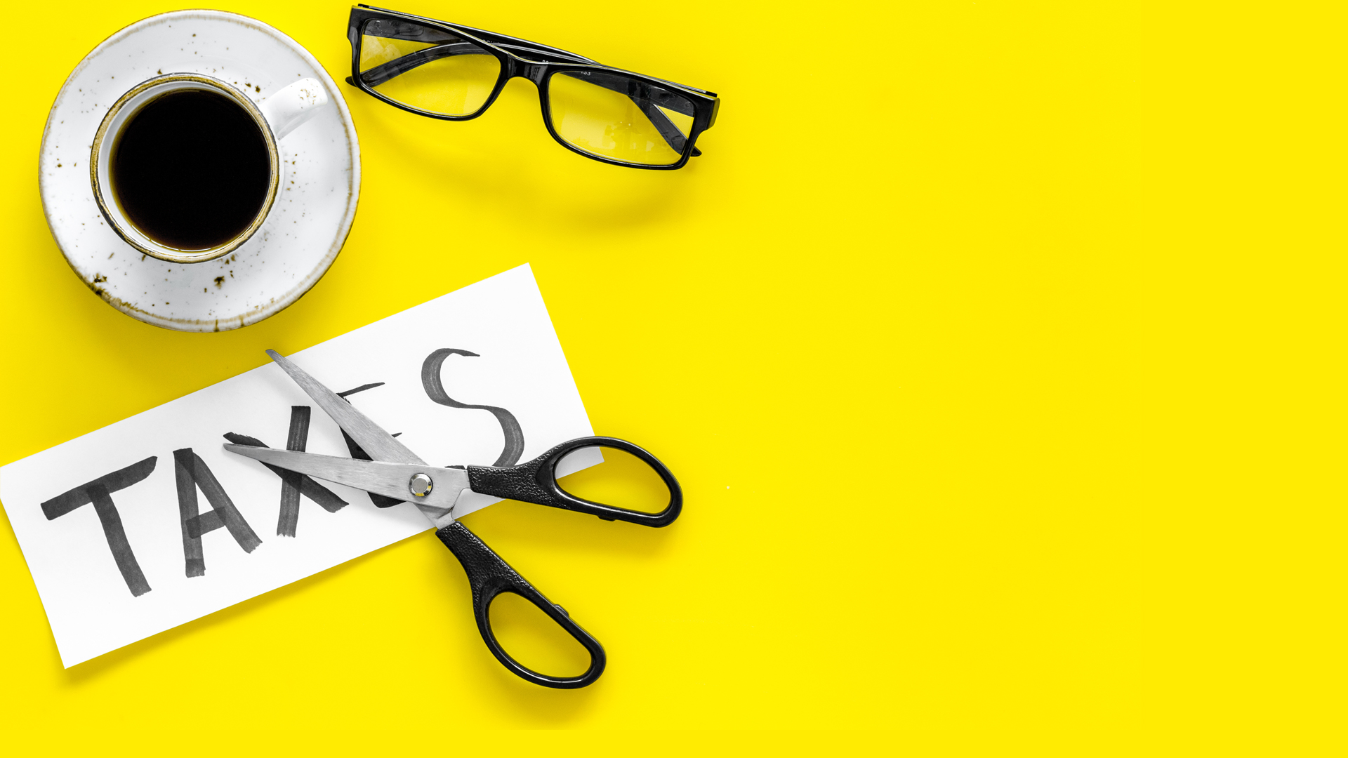 Tax reduce concept Scissors and paper with word Taxes on yellow background with coffee and glasses top view copy space