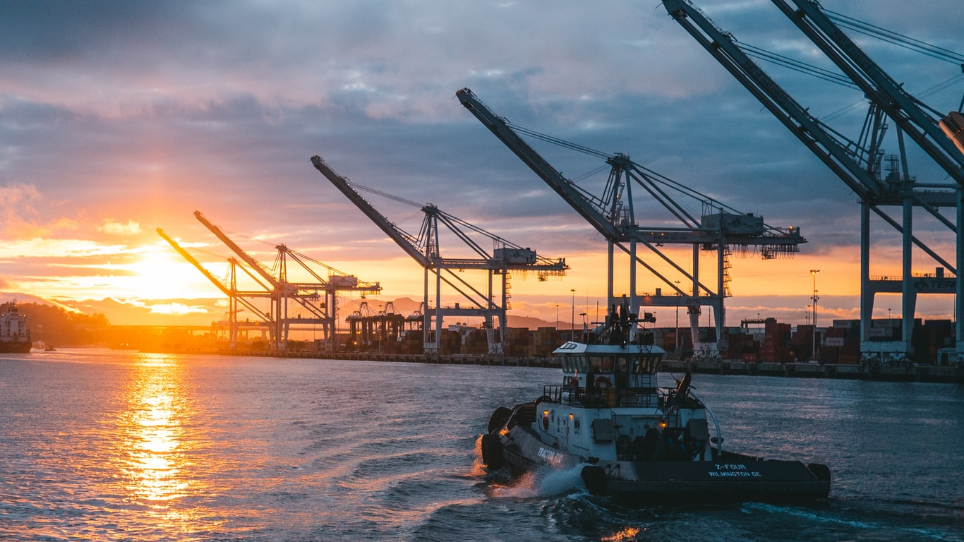 Image of a boat passing by large cranes through a canal at sunset.