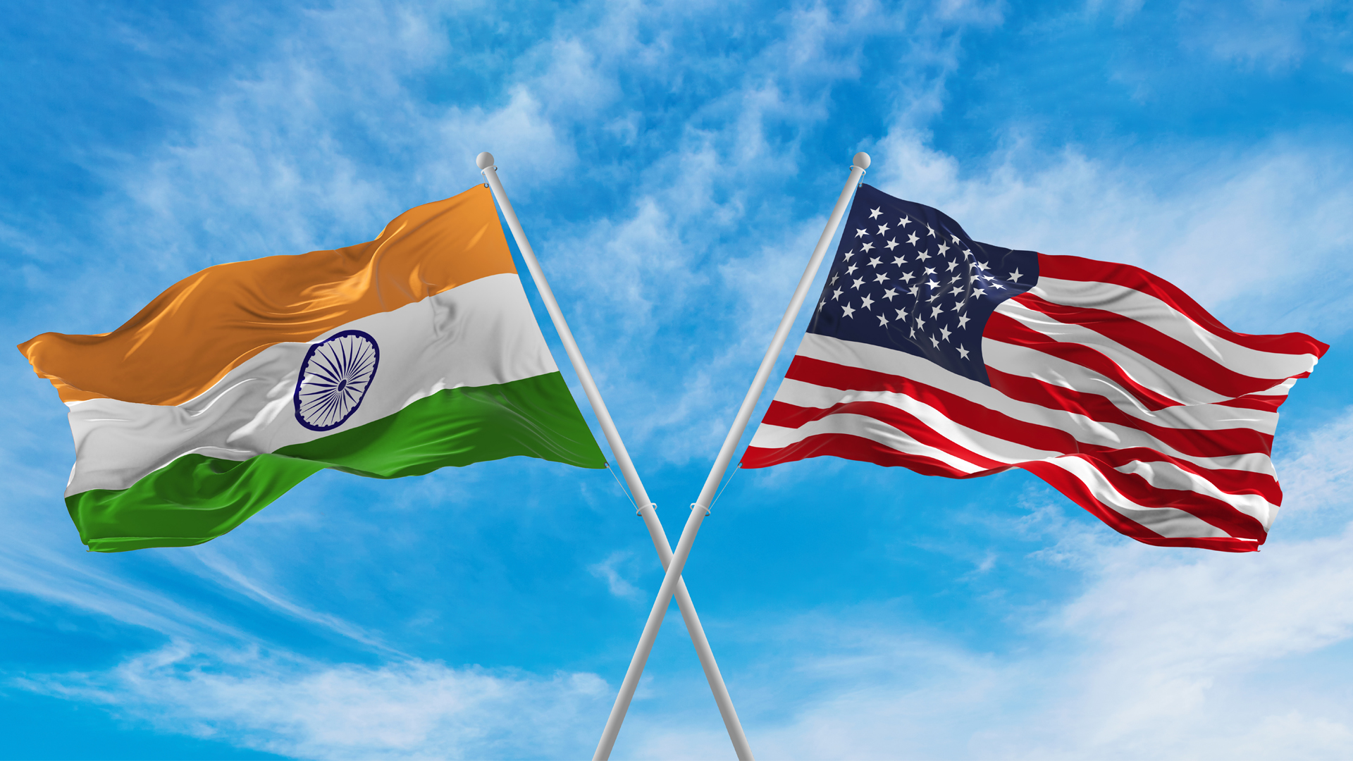 Crossed national flags of India and United States of America flag waving in the wind at cloudy blue sky.