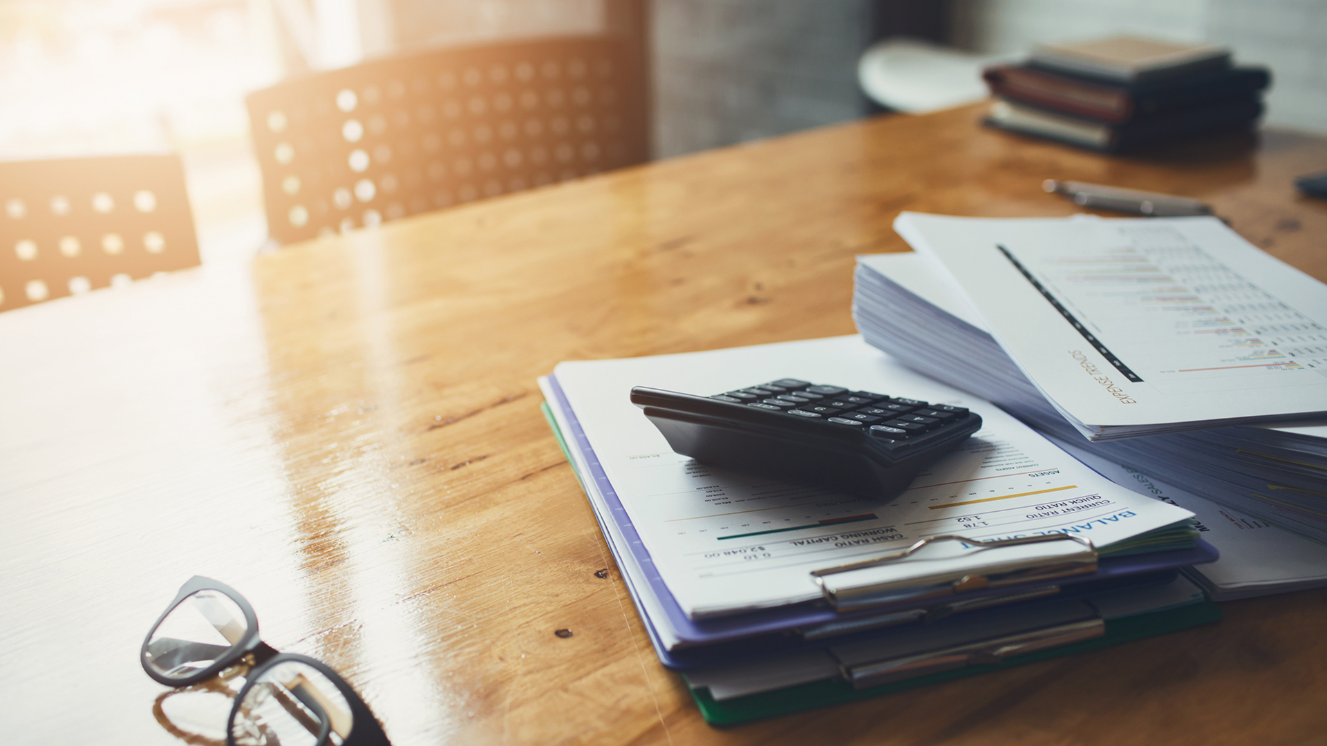 Two stacks of papers with a small black calculator sitting on a light wood table with a pair of reading glasses nearby.