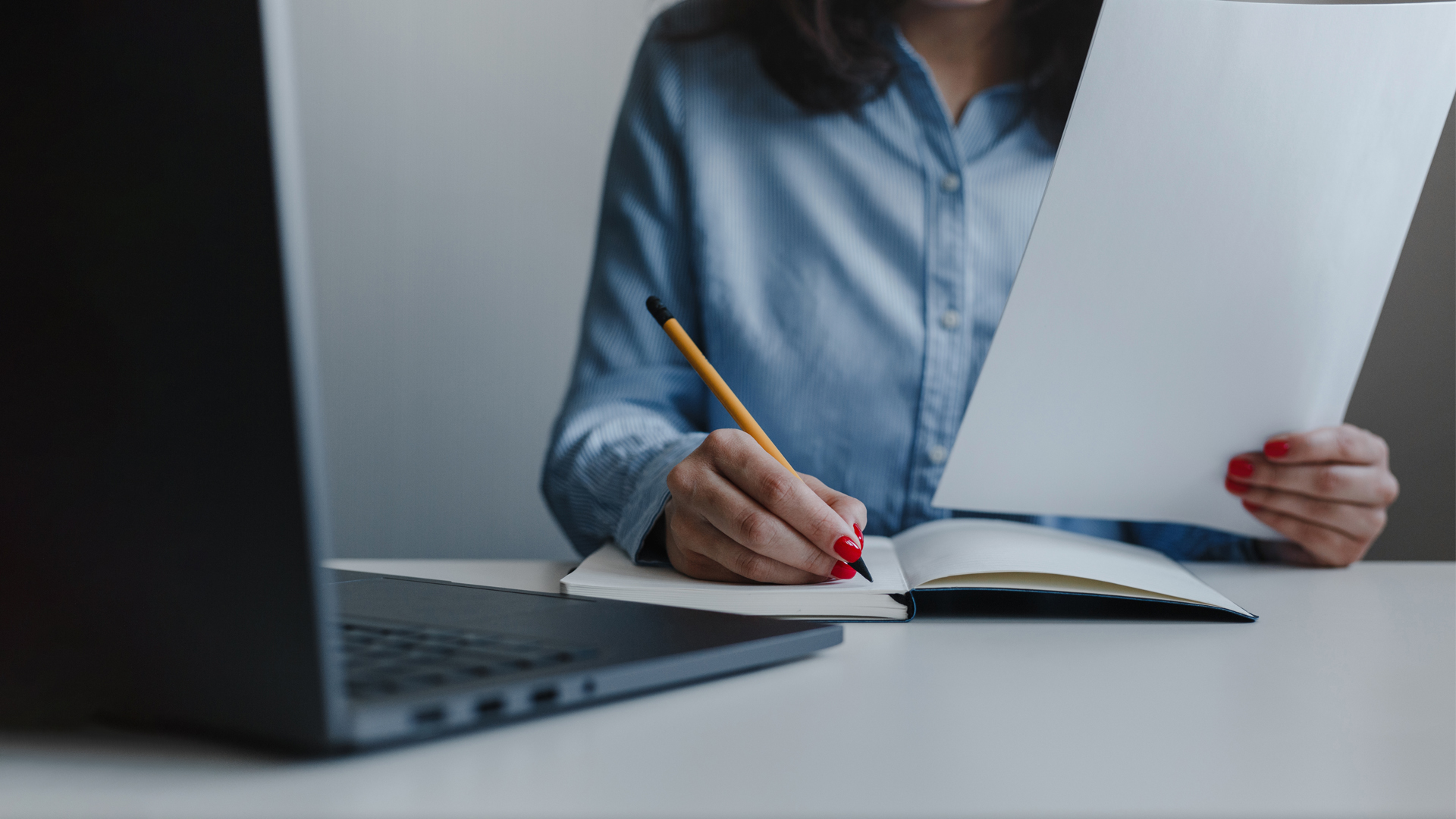 Woman wearing a light blue shirt holding a piece of paper and writing in a notebook. 