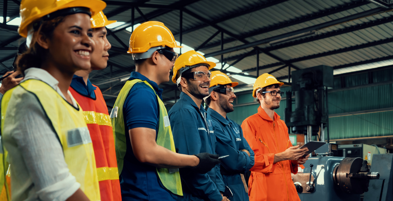 Blue collar workers stand in a line in a manufacturing facility. They are wearing hard hats and smiling.