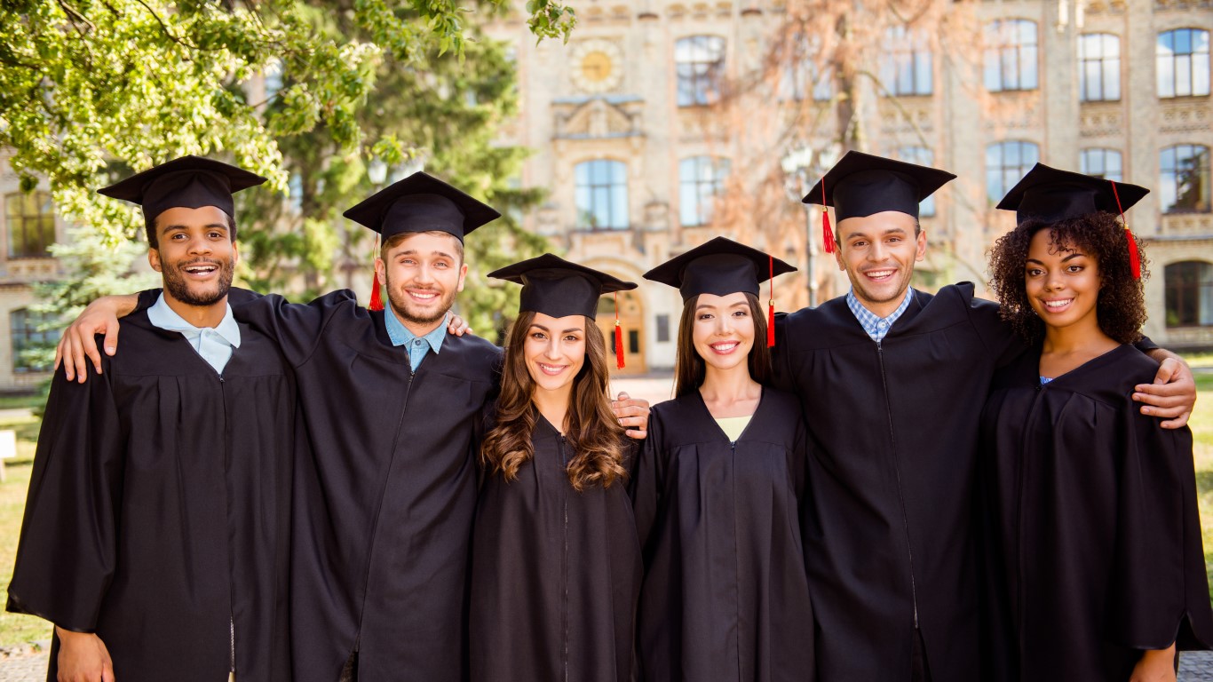 Photo of six graduates in black robes and hats smiling in front of college buildings.