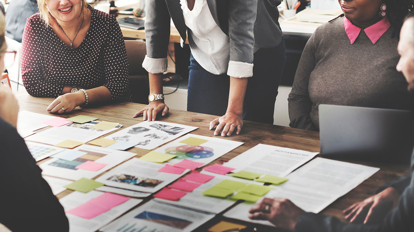 Five diverse business people huddled over a large table covered with papers and post it notes.