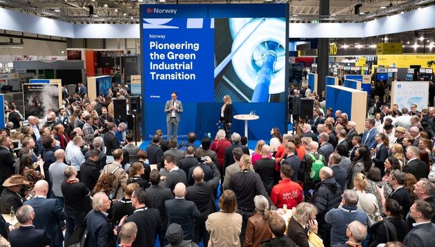 A photograph of a crowd of business people gathered around a speaker at a trade show.