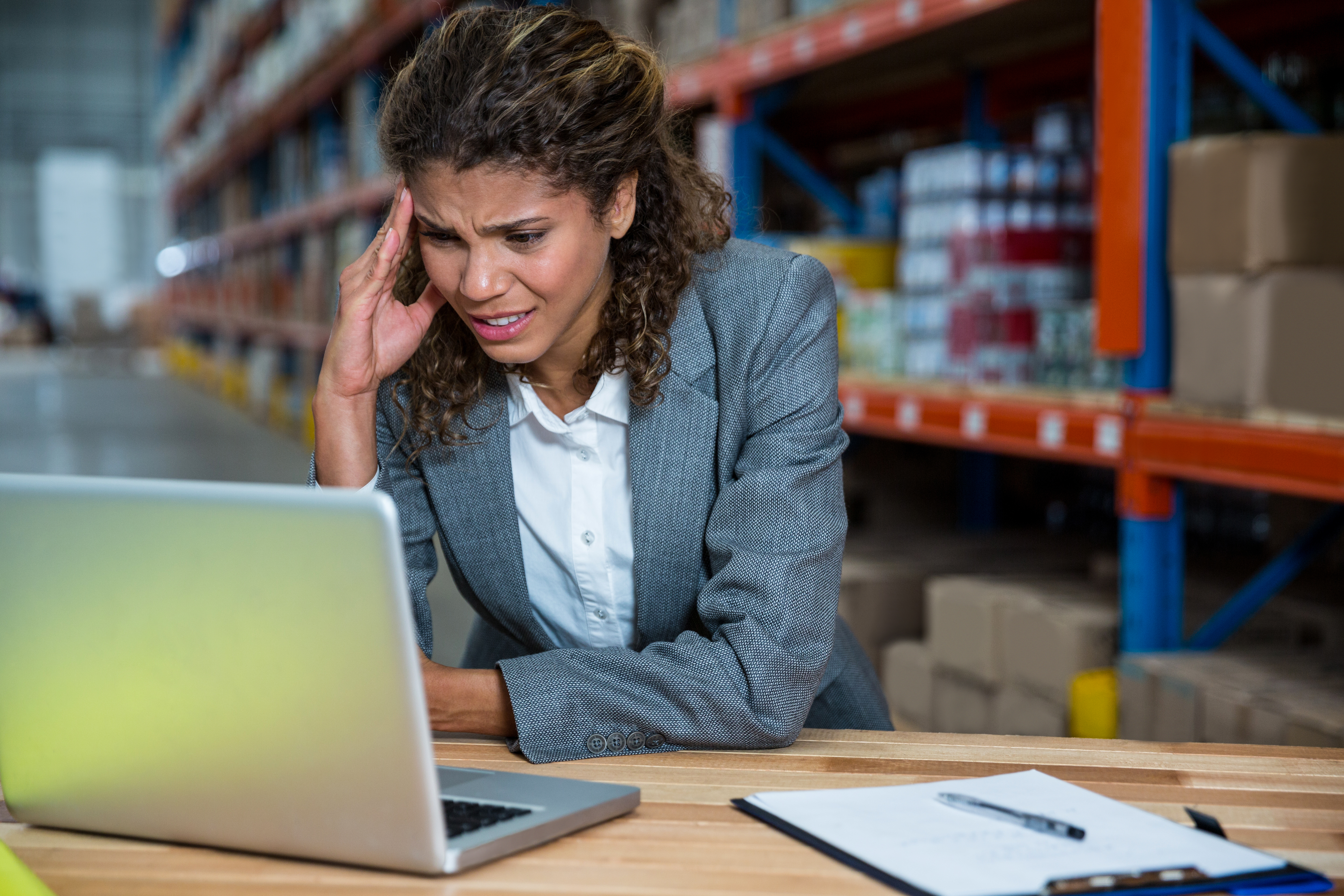 A woman in a warehouse looking at a laptop with a stressed out expression on her face.