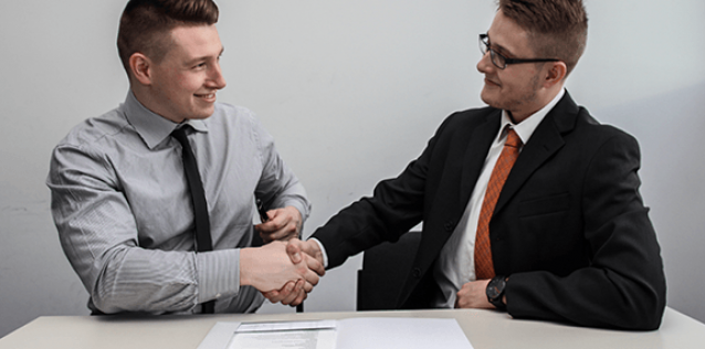 two men in business atire shaking hands while sitting at a table