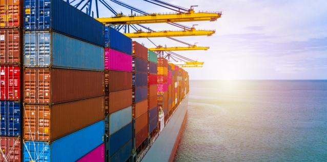 A cargo ship filled with containers in the foreground is positioned under quay cranes, with the ocean stretching out to the horizon and the sun rising and beckoning the container ship to towards its next foray across the open seas