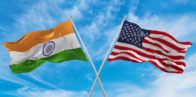 Crossed national flags of India and United States of America flag waving in the wind at cloudy blue sky.