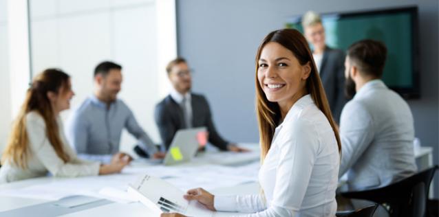 Woman looking back at the camera smiling and sitting next to a group of business people collaborating in office.