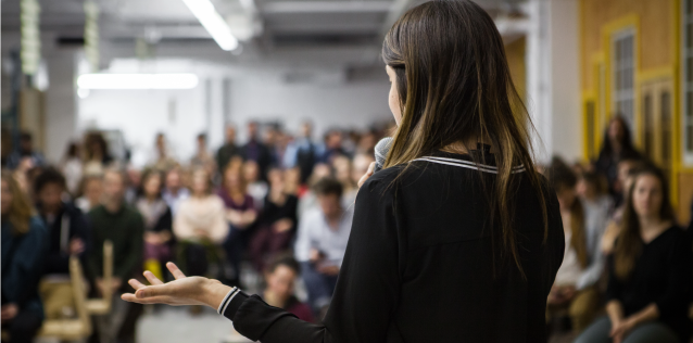 Photo of a woman giving a presentation to a room full of people. 