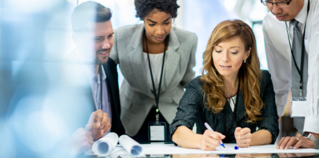 Four professionals looking at a white paper and a woman writing text on a paper and the other men and woman looking down.