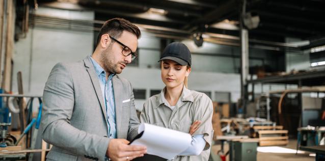 An inspector in a suit is holding up a file and inquiring about it to a female factory worker.
