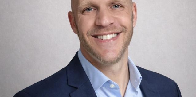 A professional headshot of a smiling man with short light brown hair and trimmed facial hair, wearing a navy blazer over a light blue dress shirt.