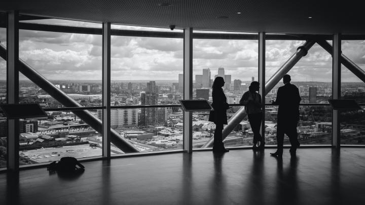 There persons having a discussion in front of a large window looking out on a city skyline