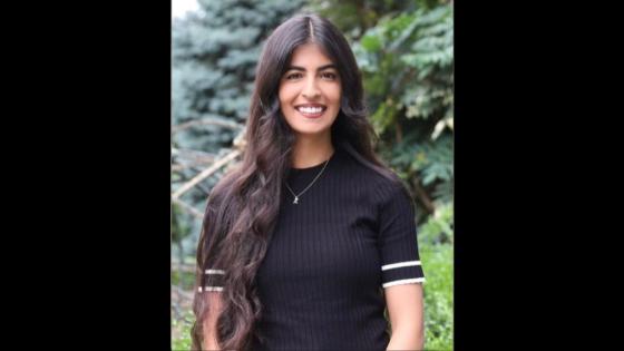 Woman with long black hair wearing a black shirt standing in front of green trees and bushes