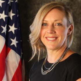 Bust photo of Frances Selema beside a draped American Flag wearing a black knit top and silver jewelry, .