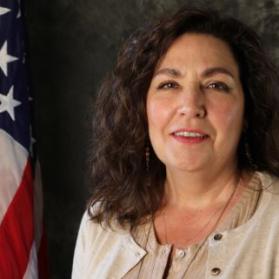 Bust photo of Juanita Harthun beside an American flag, wearing a tan cardigan over a tan knit top.