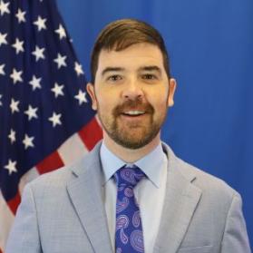 Bust photo of Justin Tapp in front in of a draped American Flag wearing a gray blazer, white collared shirt, and blue patterned tie.